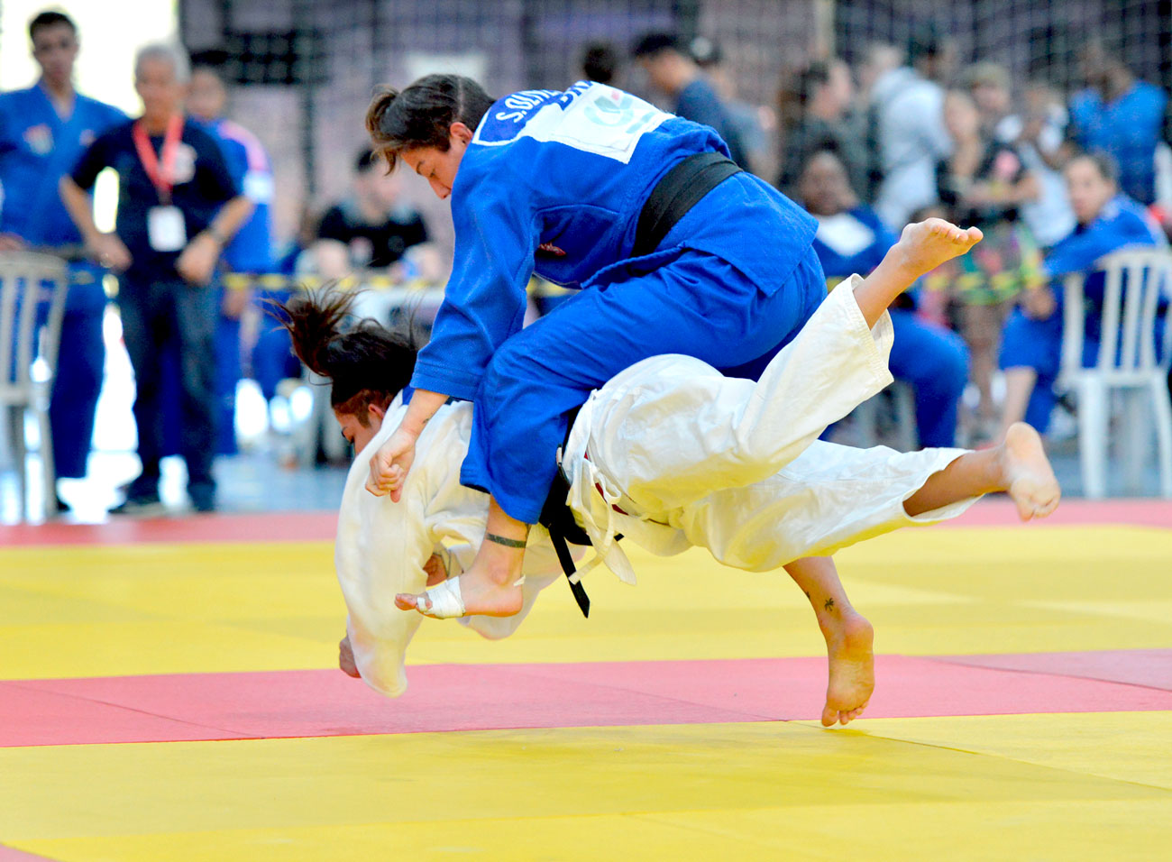 Uma mulher demonstrando suas habilidades com uniforme de judô azul e amarelo em uma competição inter-regional.