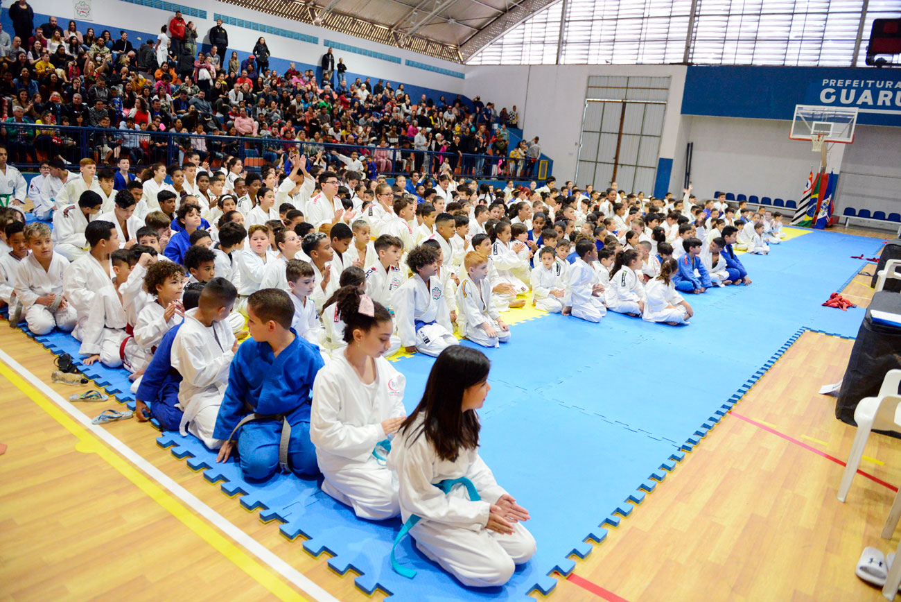 Um grupo de crianças uniformizadas de caratê sentadas em uma academia, homenageia o sensei Miguel Suganuma.