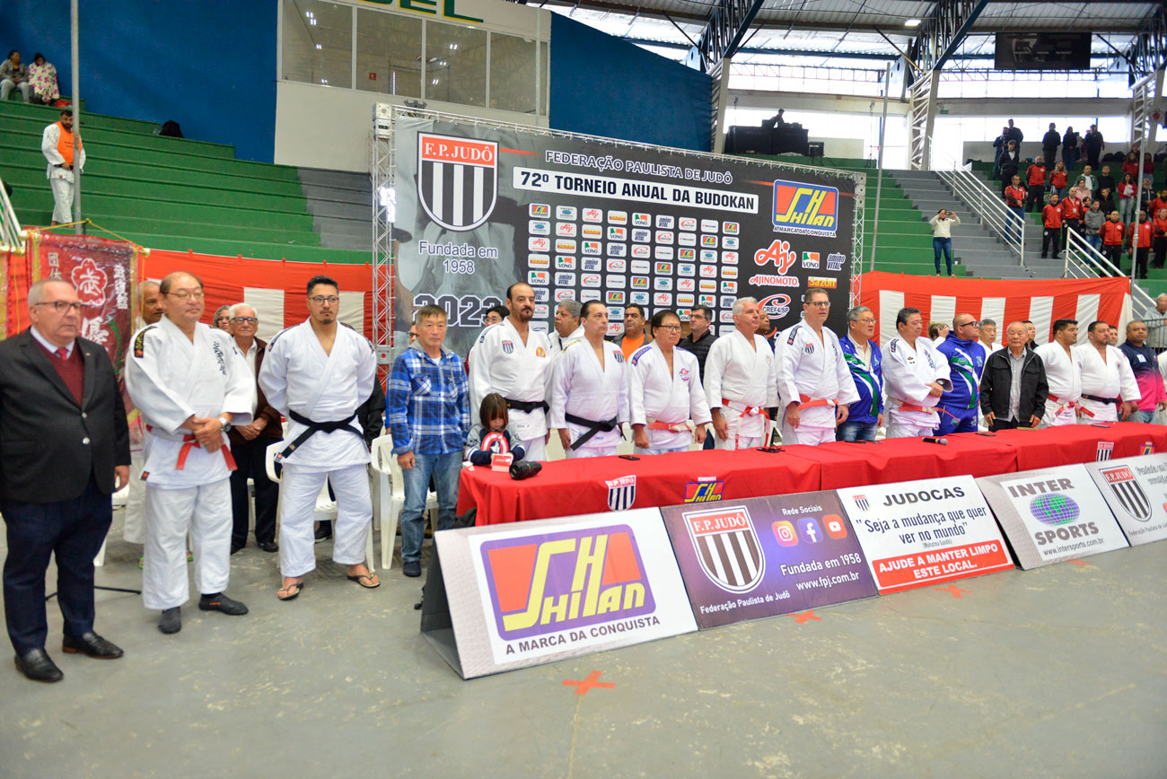 Um grupo de pessoas posando para foto em frente a um banner histórico na 72ª edição do Torneio Budokan.